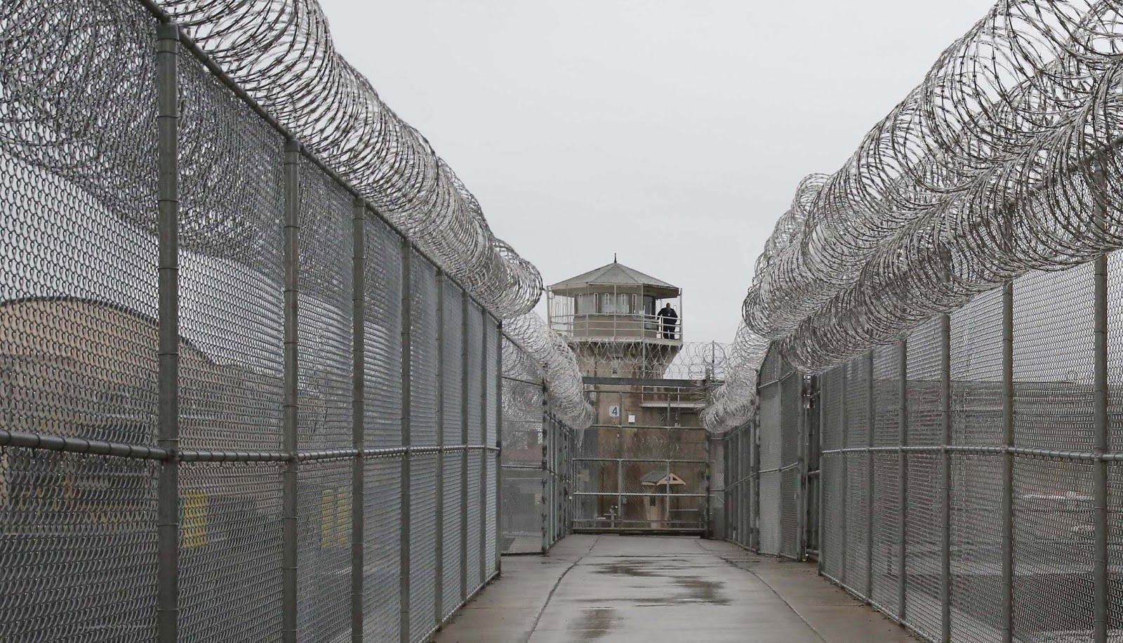 A colored photograph of Washington State Penitentiary’s watch tower from the outside, as seen down a concrete corridor. High fences topped with barbed wire line each side of the corridor.