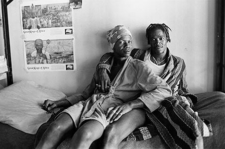 A black and white photo of two people sitting together, against a wall and on top of a cell bed. There is a poster above the bed. Kim’s arm is wrapped around Leomy’s shoulder, and Leomy’s body is nestled into Kim’s. Both are wearing different colored robes and are looking at the camera.