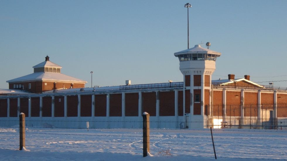 A wintertime image of the Saskatchewan Penitentiary. The brown walls are interspersed with white columns, and there are two towers at either end of the building. A barbed wire fence surrounds the property.