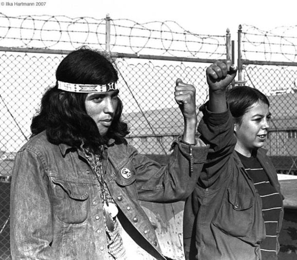 An Indigenous man and woman stand with fists raised in front of a barbed wire fence. They are involved in the occupation of Alcatraz Island, one of the many actions of the American Indian Movement.