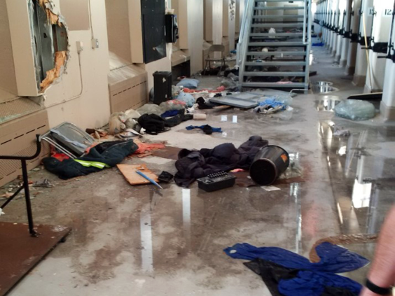 Aftermath of the 2016 Riot at the Saskatchewan Penitentiary. A hallway with garbage and water is flanked on the right-hand side by a row of cell doors. On the left-hand side, drywall has been pulled off a protruding portion of the wall.