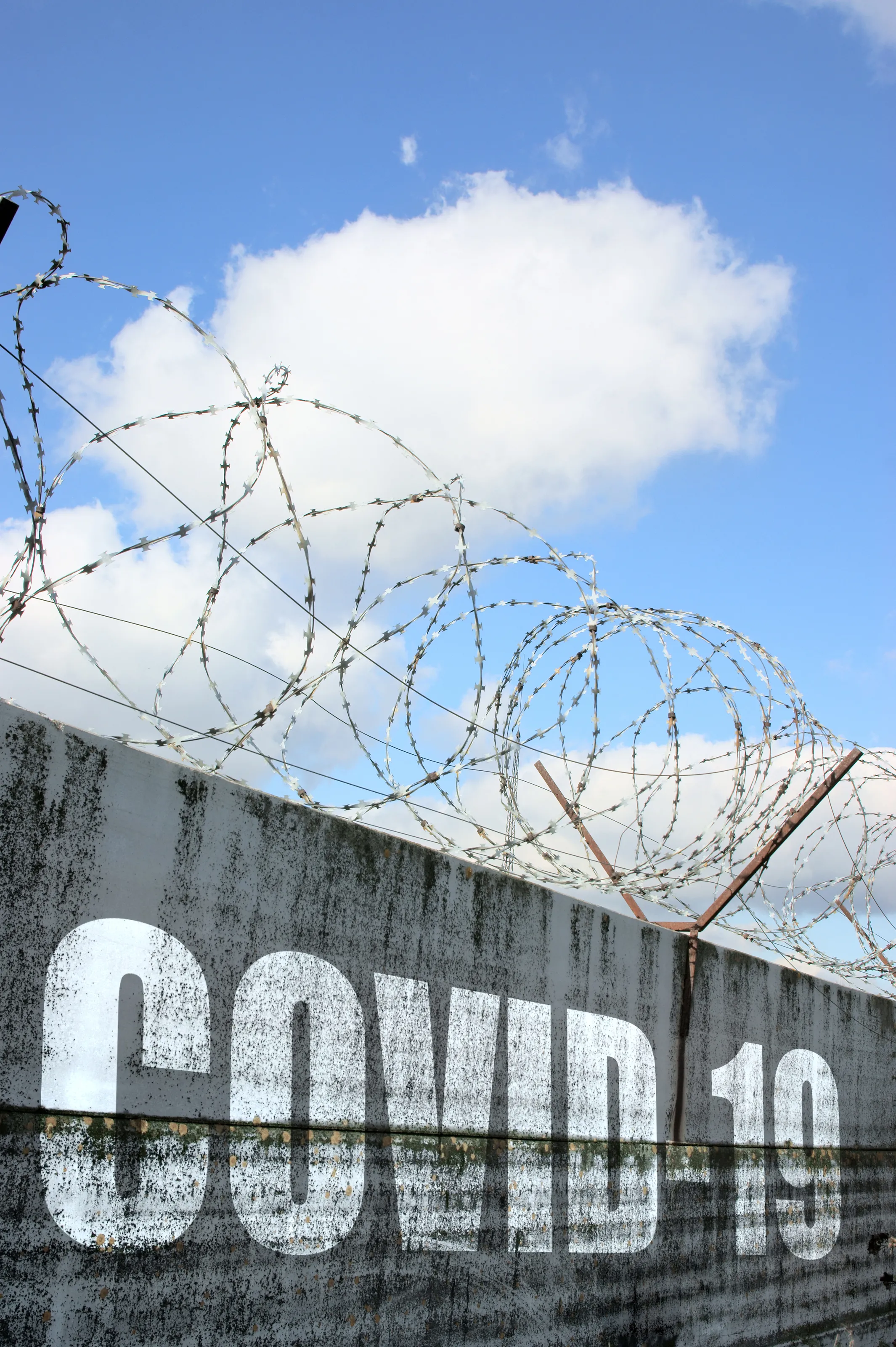 A concrete wall with “COVID-19” in big letters. Barbed wire is at the top of the wall.